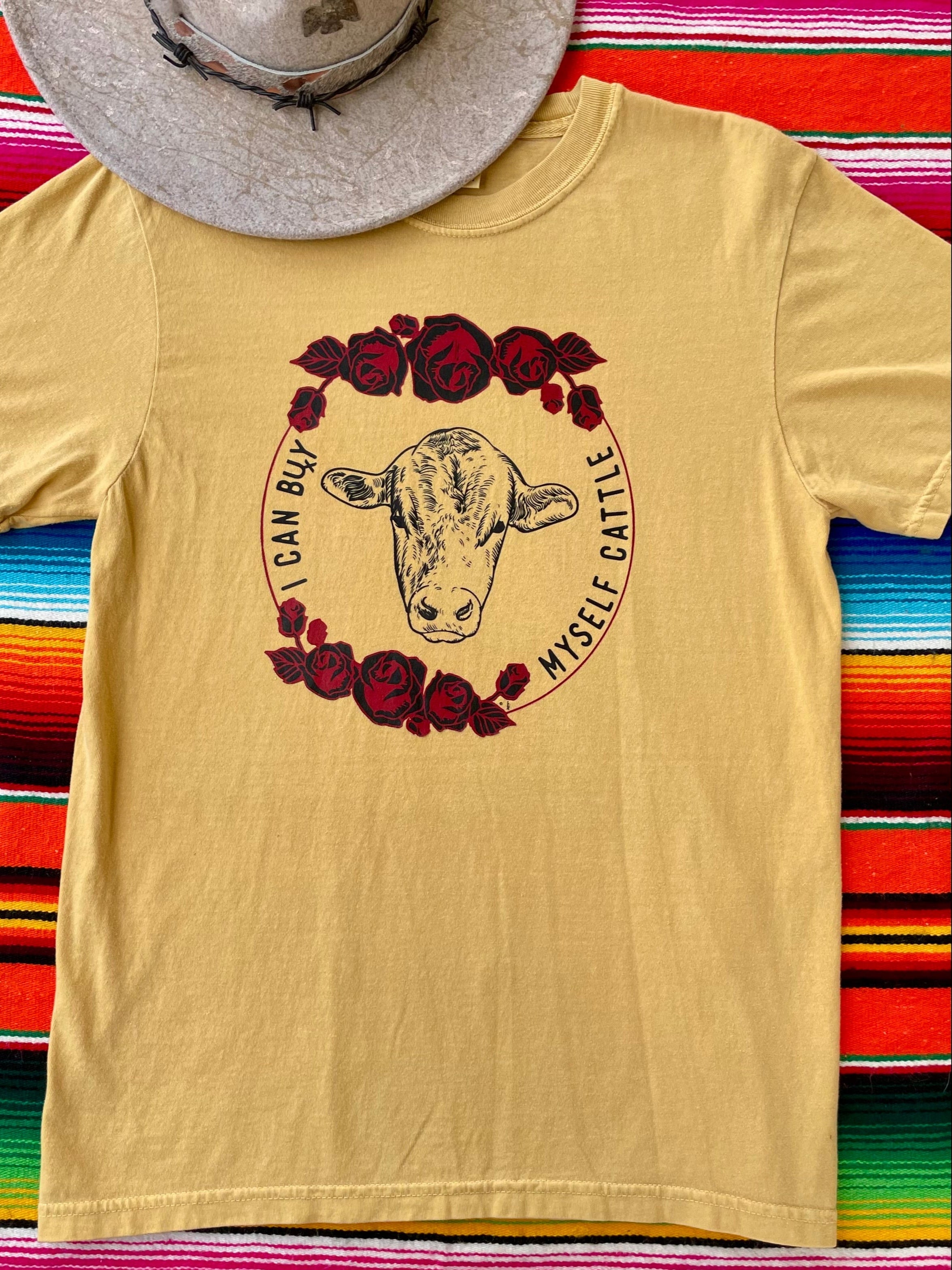 Mustard Yellow western graphic tee with cow head and red roses, bold slogan “Can Buy Myself Cattle” inspired by Miley Cyrus. Feminine cowgirl shirt with country sass and southern style. Rodeo-ready and perfect for bold fashion lovers.
