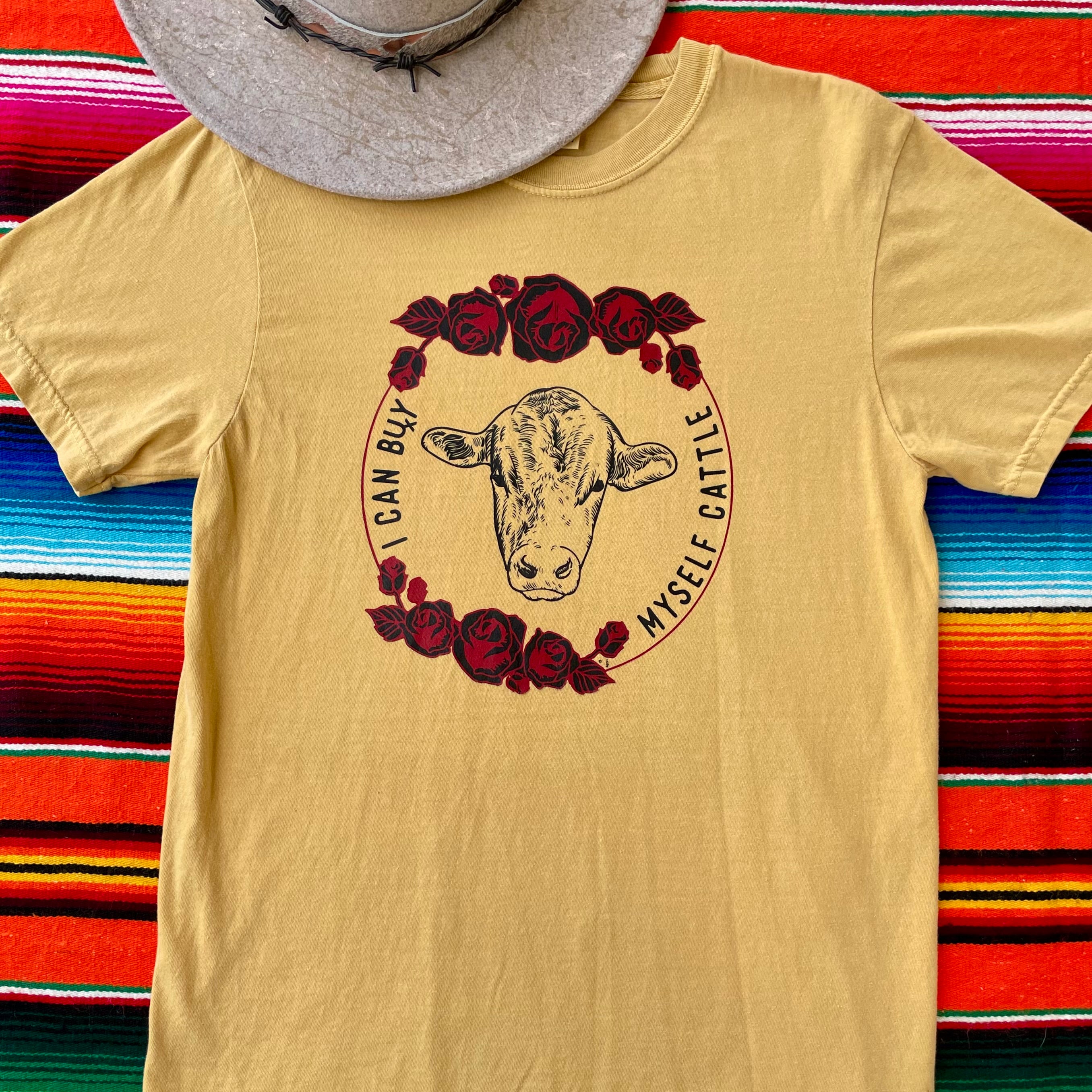 Mustard Yellow western graphic tee with cow head and red roses, bold slogan “Can Buy Myself Cattle” inspired by Miley Cyrus. Feminine cowgirl shirt with country sass and southern style. Rodeo-ready and perfect for bold fashion lovers.