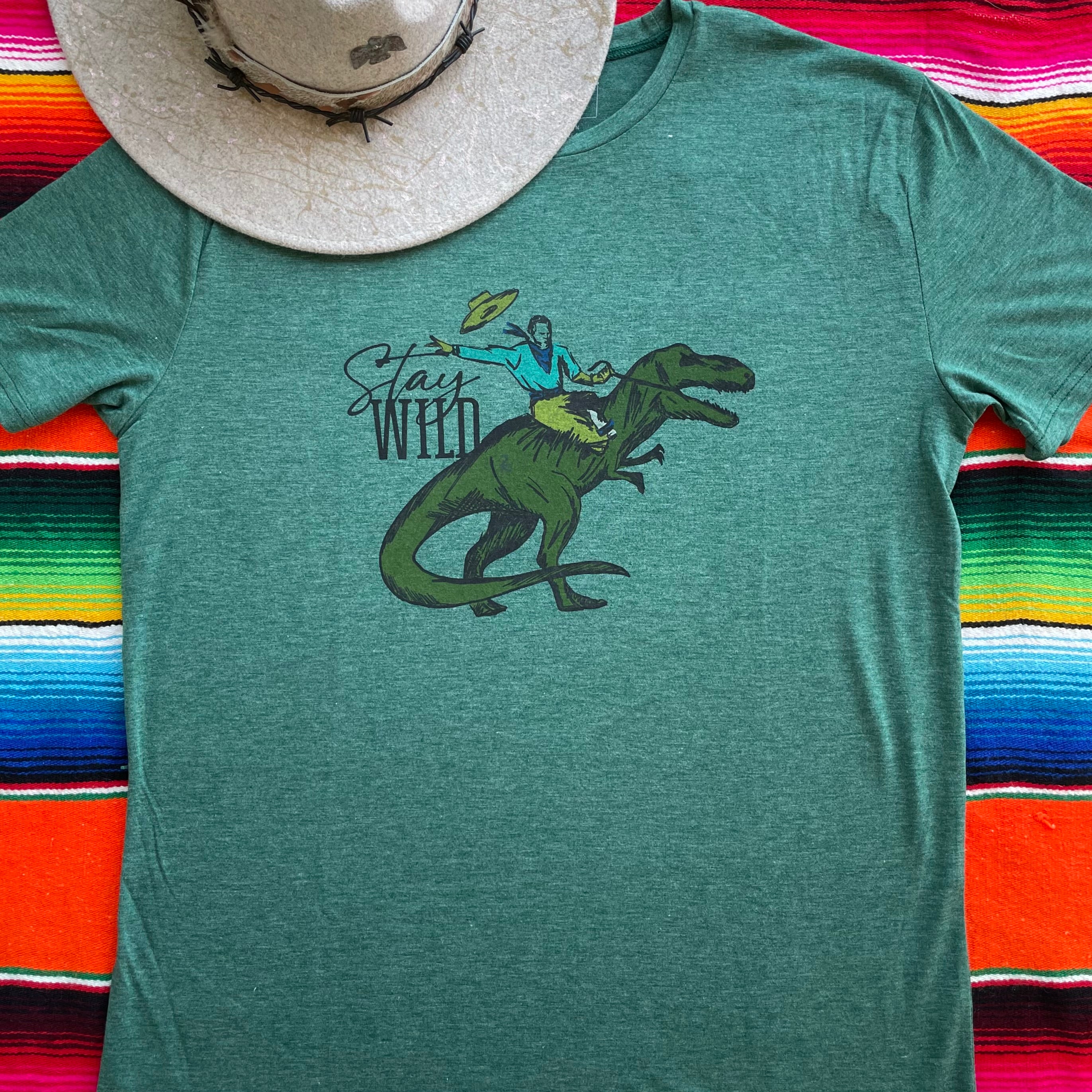 Green t-shirt with a graphic of a dinosaur and text 'Stay Weird' on a colorful striped background