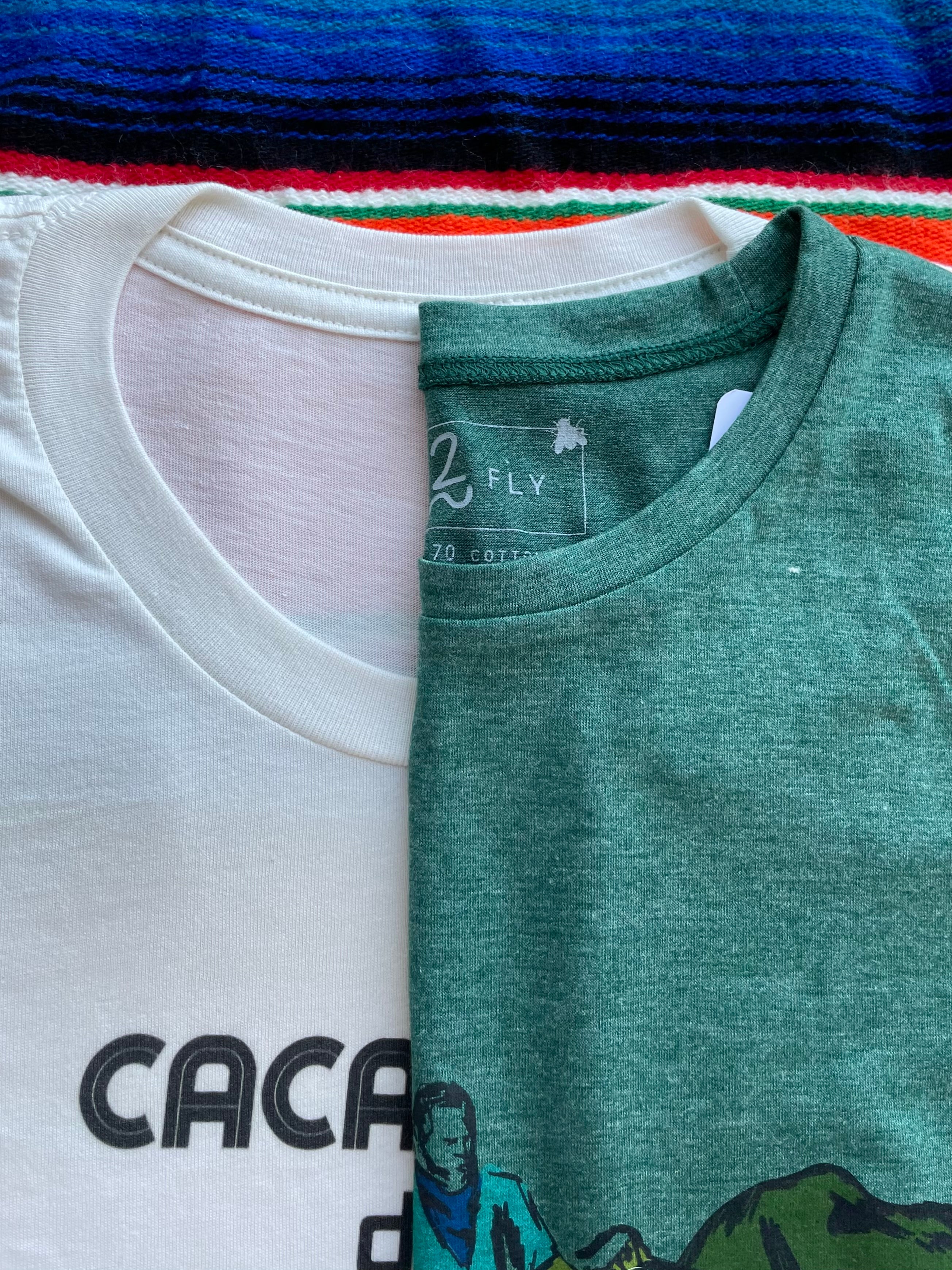 Two folded t-shirts, one white with 'Cacao' text and the other green with a graphic design, on a colorful background.