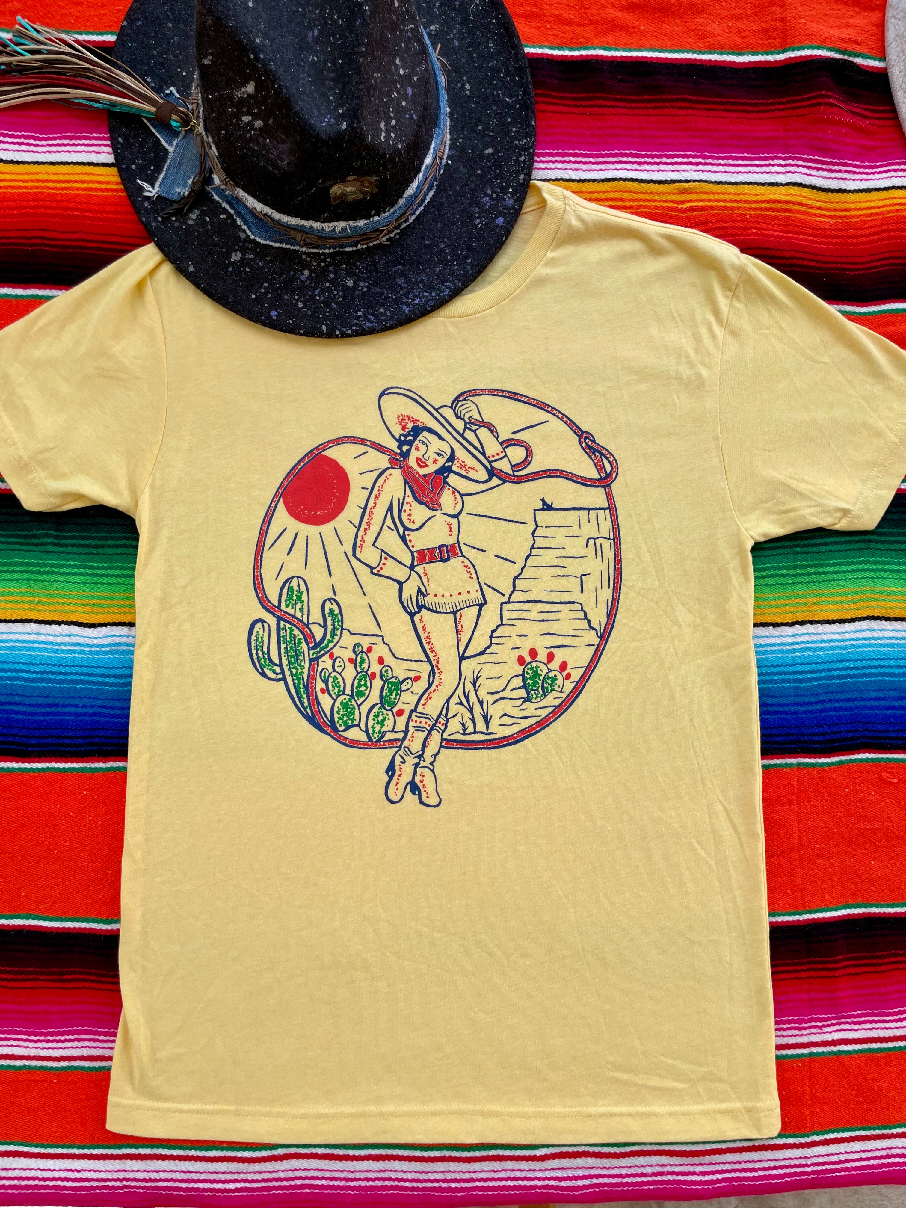 Yellow t-shirt with a graphic design of a woman in a desert scene on a colorful striped background.