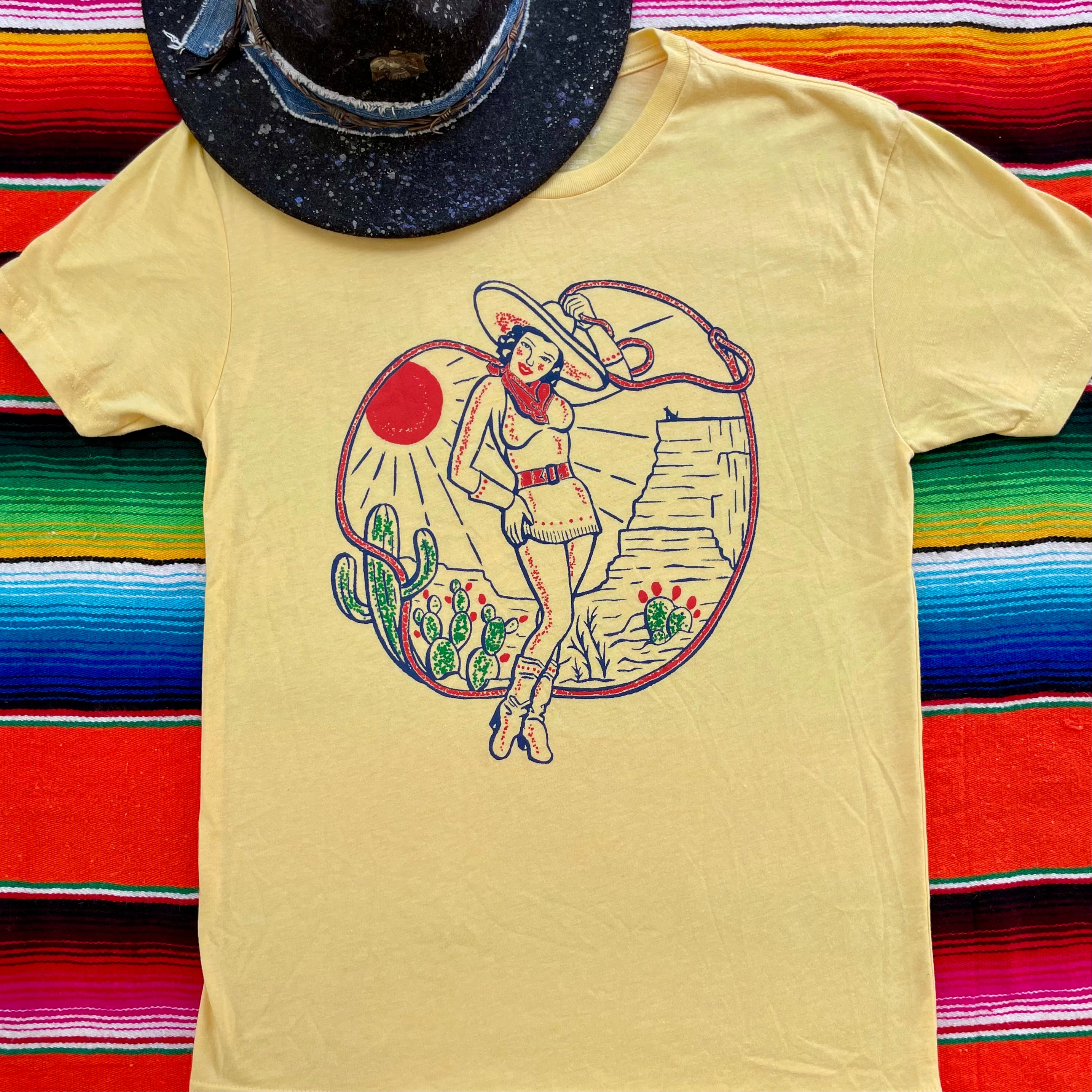 Yellow t-shirt with a graphic design of a woman in a hat and skirt, cactus, and sun on a colorful striped background.