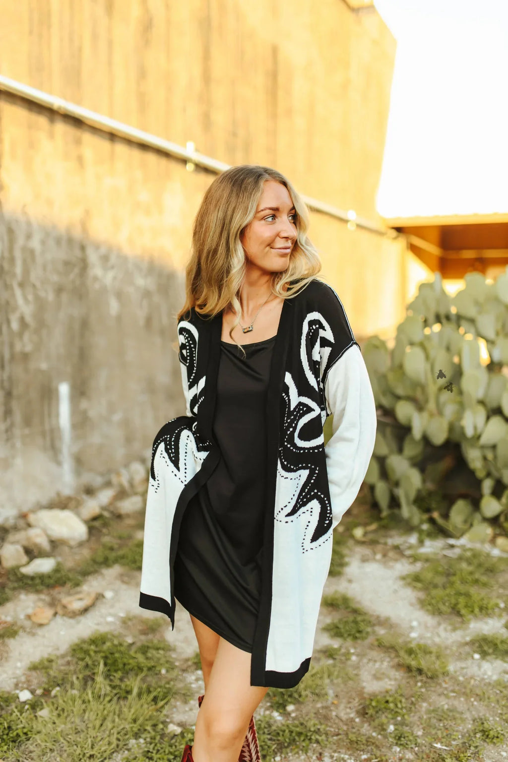 Woman wearing a black and white patterned cardigan over a black dress, standing outdoors with a cactus in the background.