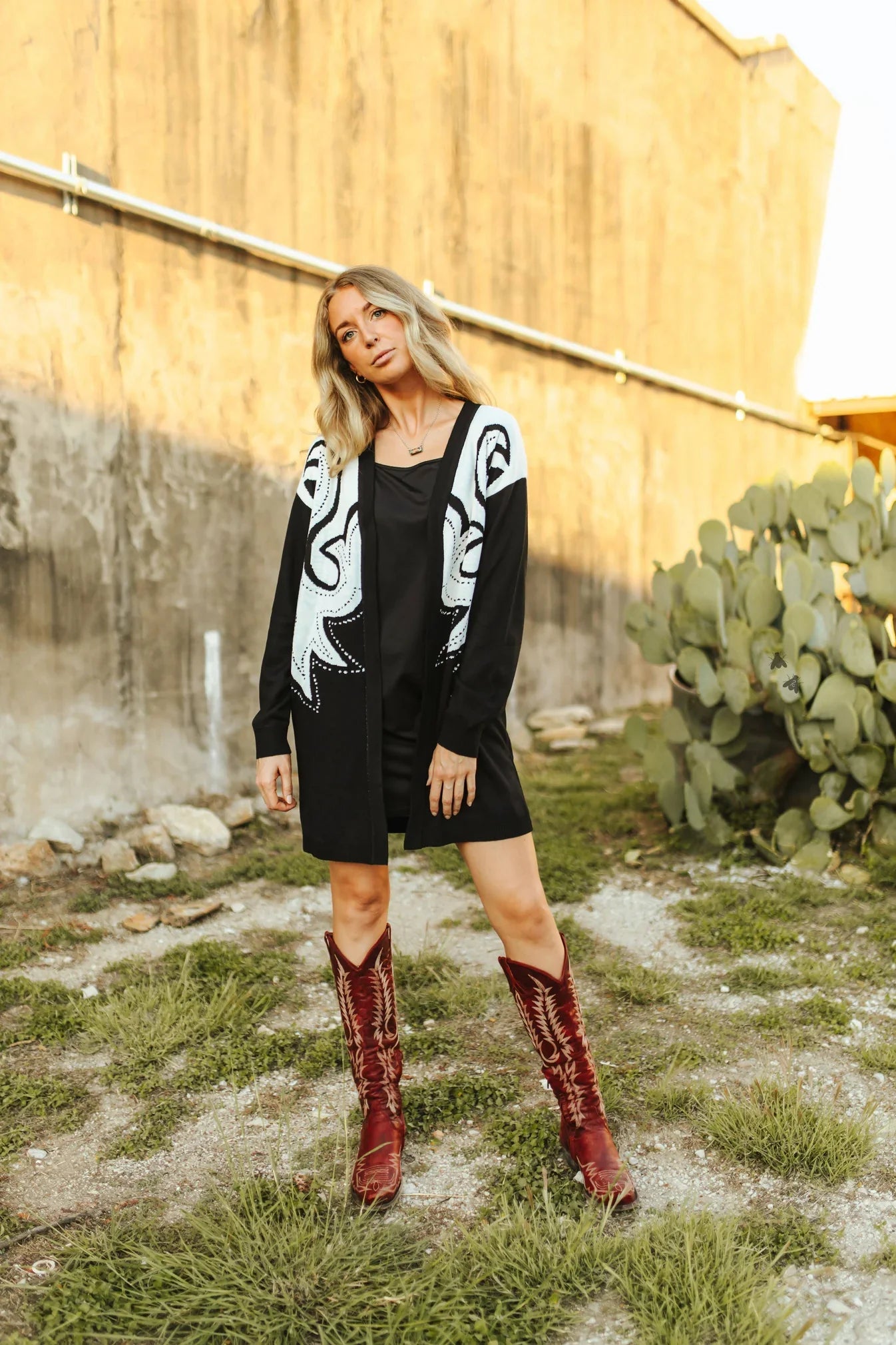 Woman wearing a black dress with white patterns, red boots, and a black cardigan with white designs outdoors.