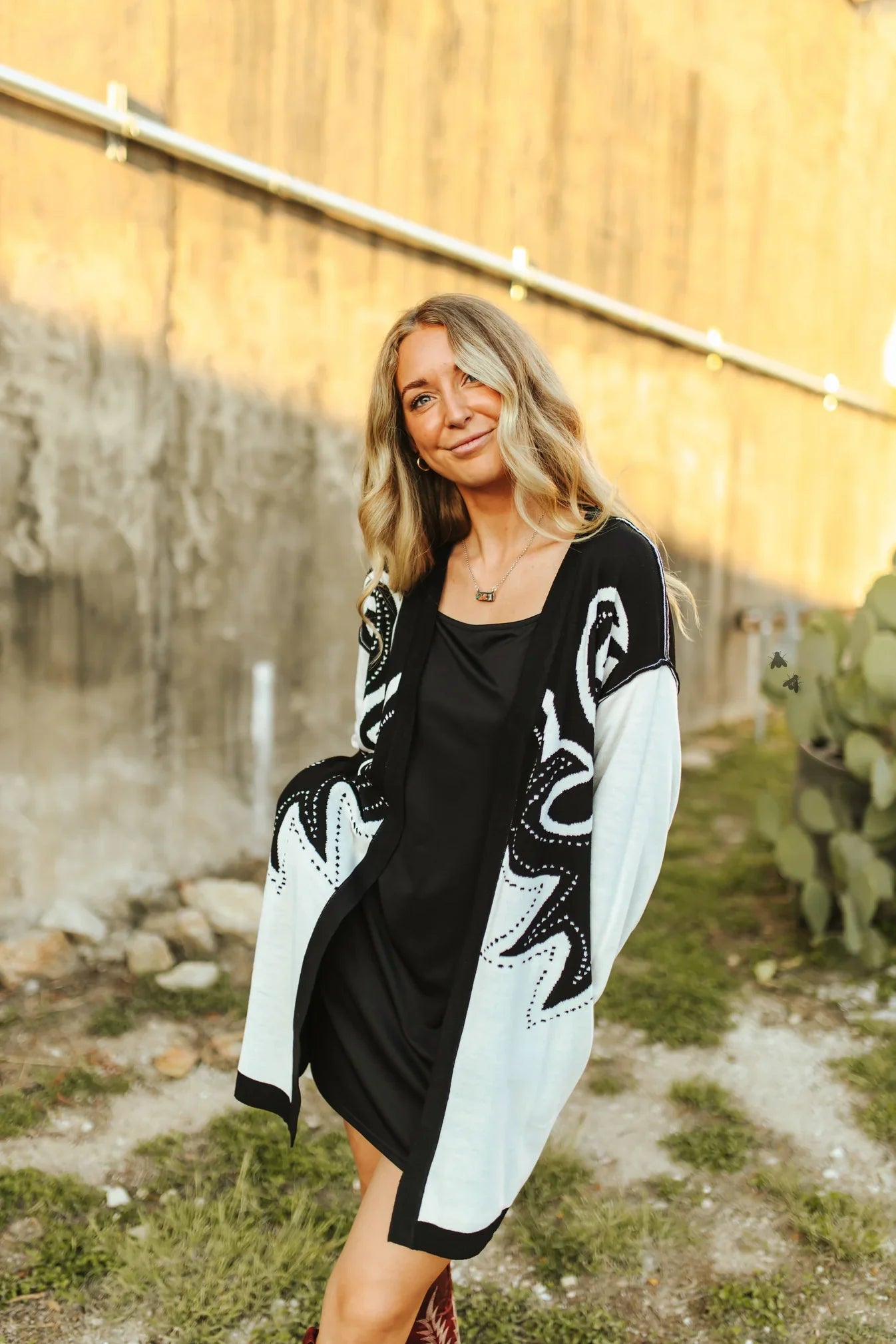 Woman wearing a black and white patterned cardigan outdoors with a wooden fence and cactus in the background.