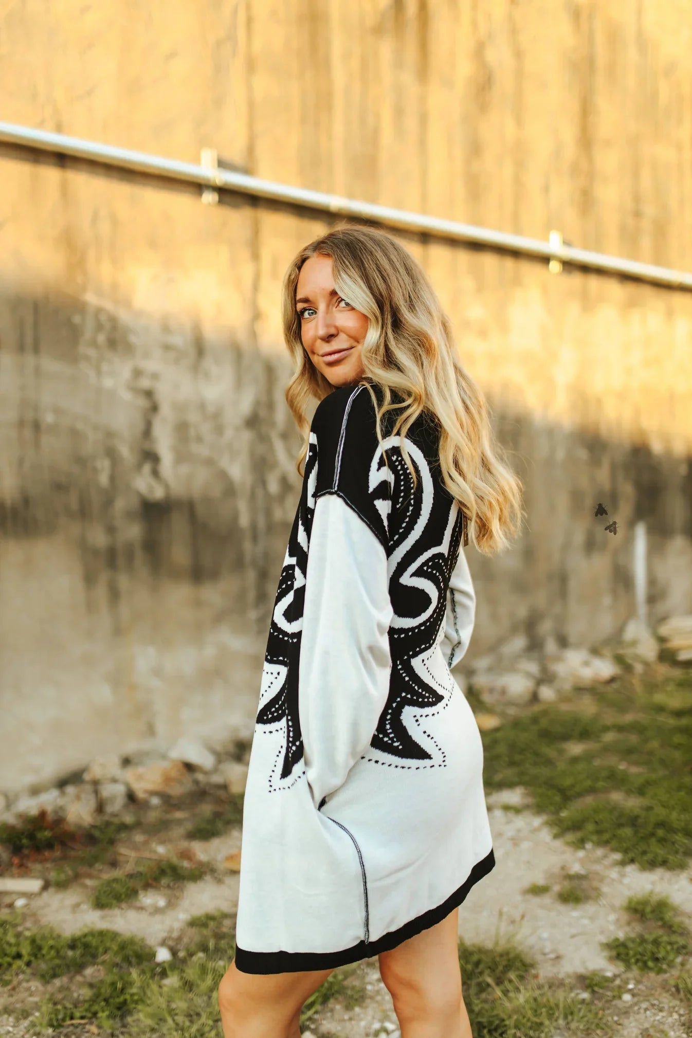 Woman wearing a white dress with black patterns standing outdoors near a wall.