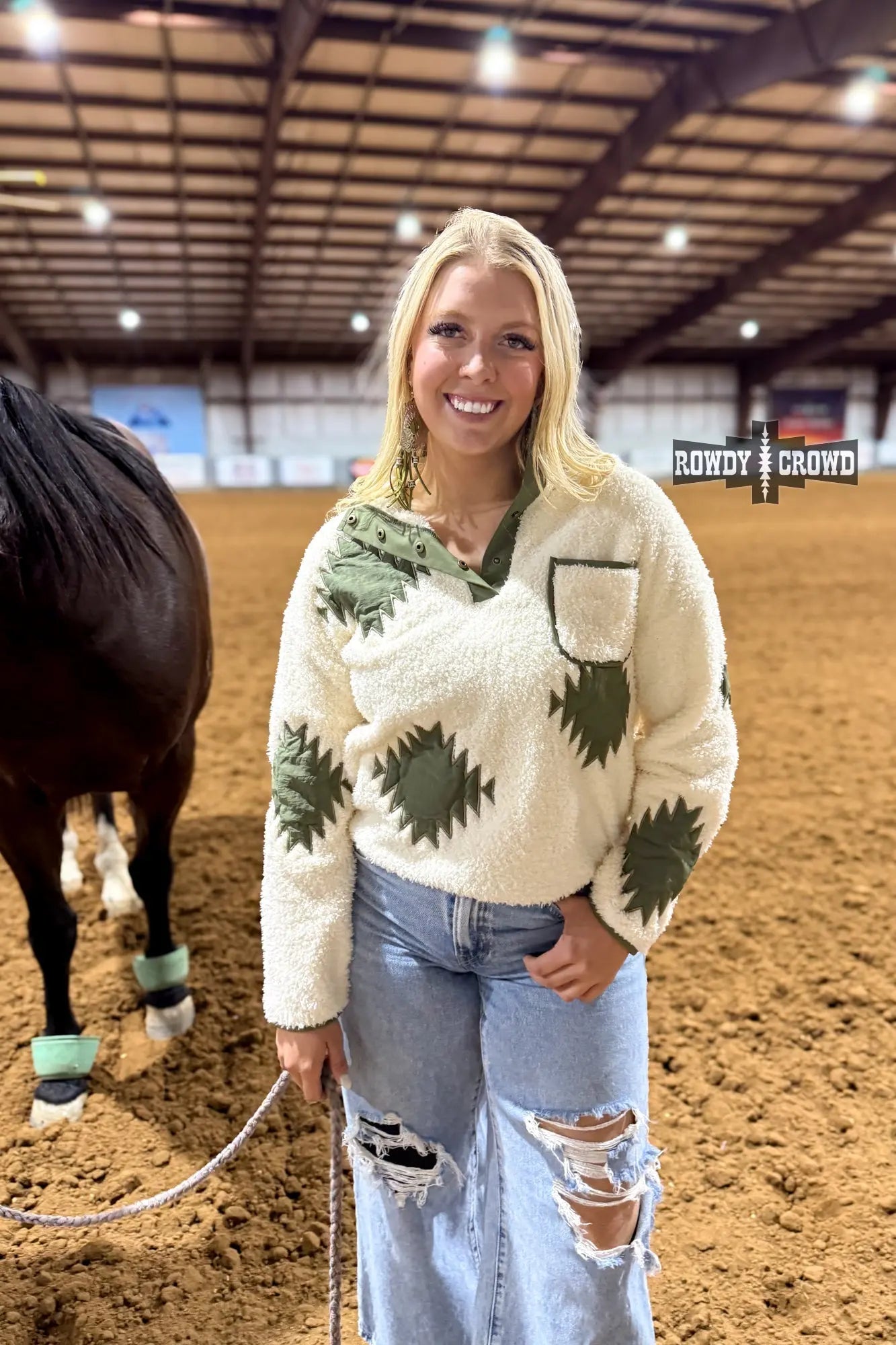 Woman wearing cream western sherpa pullover with olive Aztec patches, snap‑button neckline, and ripped jeans, standing in equestrian arena.